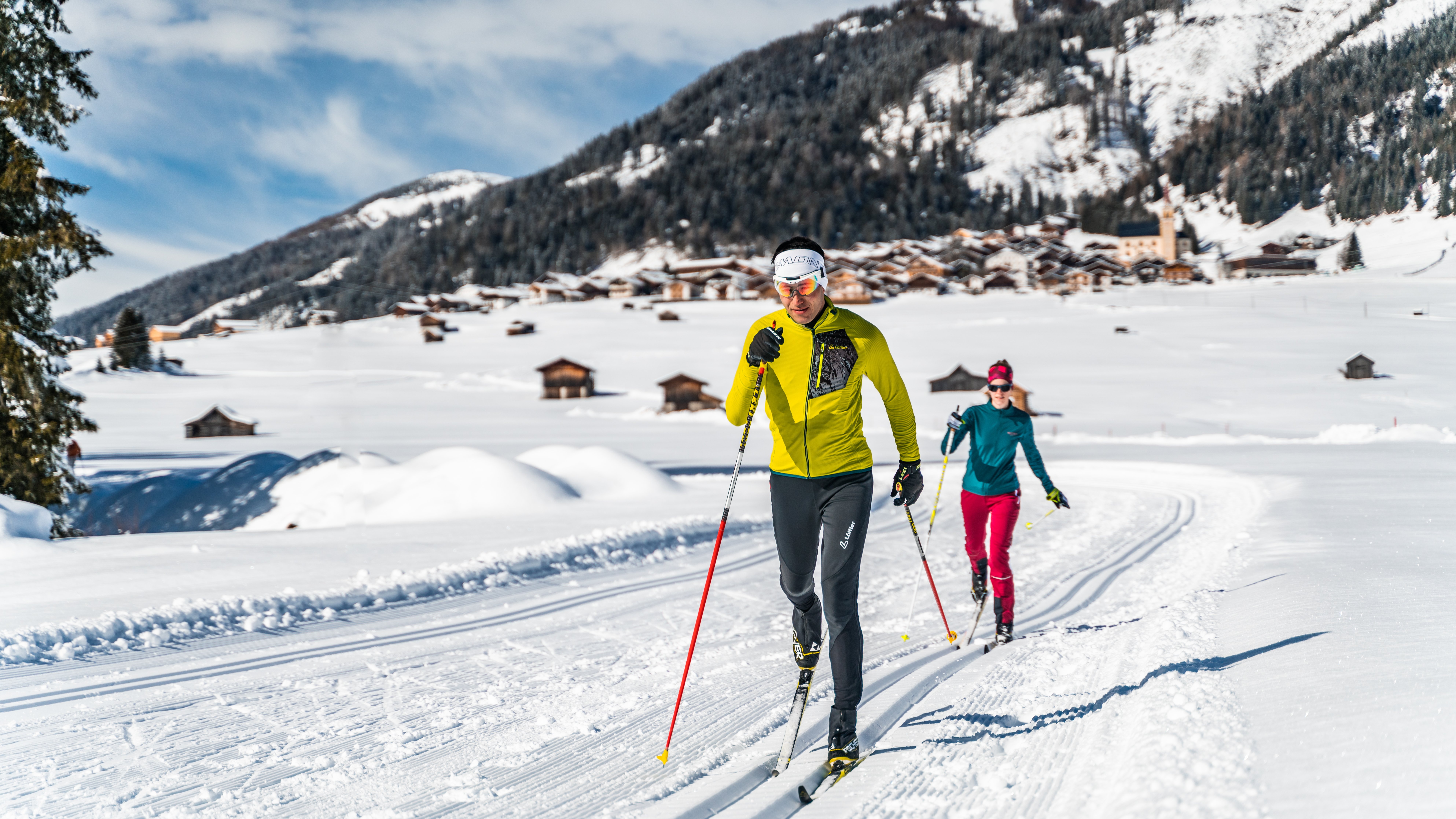 Two people cross-country skiing on a groomed trail in a snow-covered mountain landscape under blue skies. ©TVB Osttirol Peter Maier Obertilliach