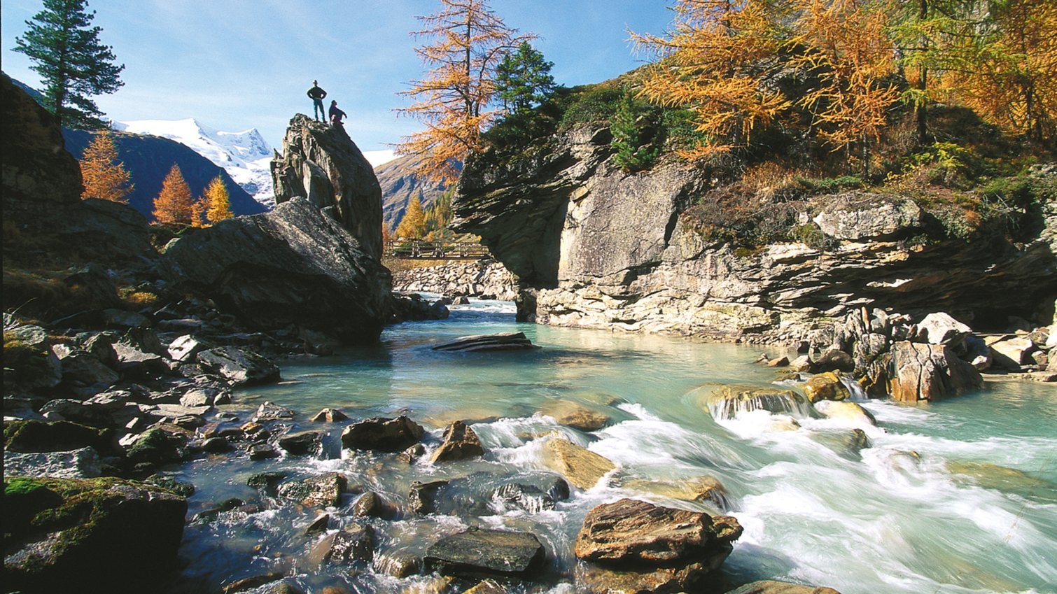 Zwei Personen auf Felsen über einem klaren Gebirgsbach in herbstlicher Landschaft.