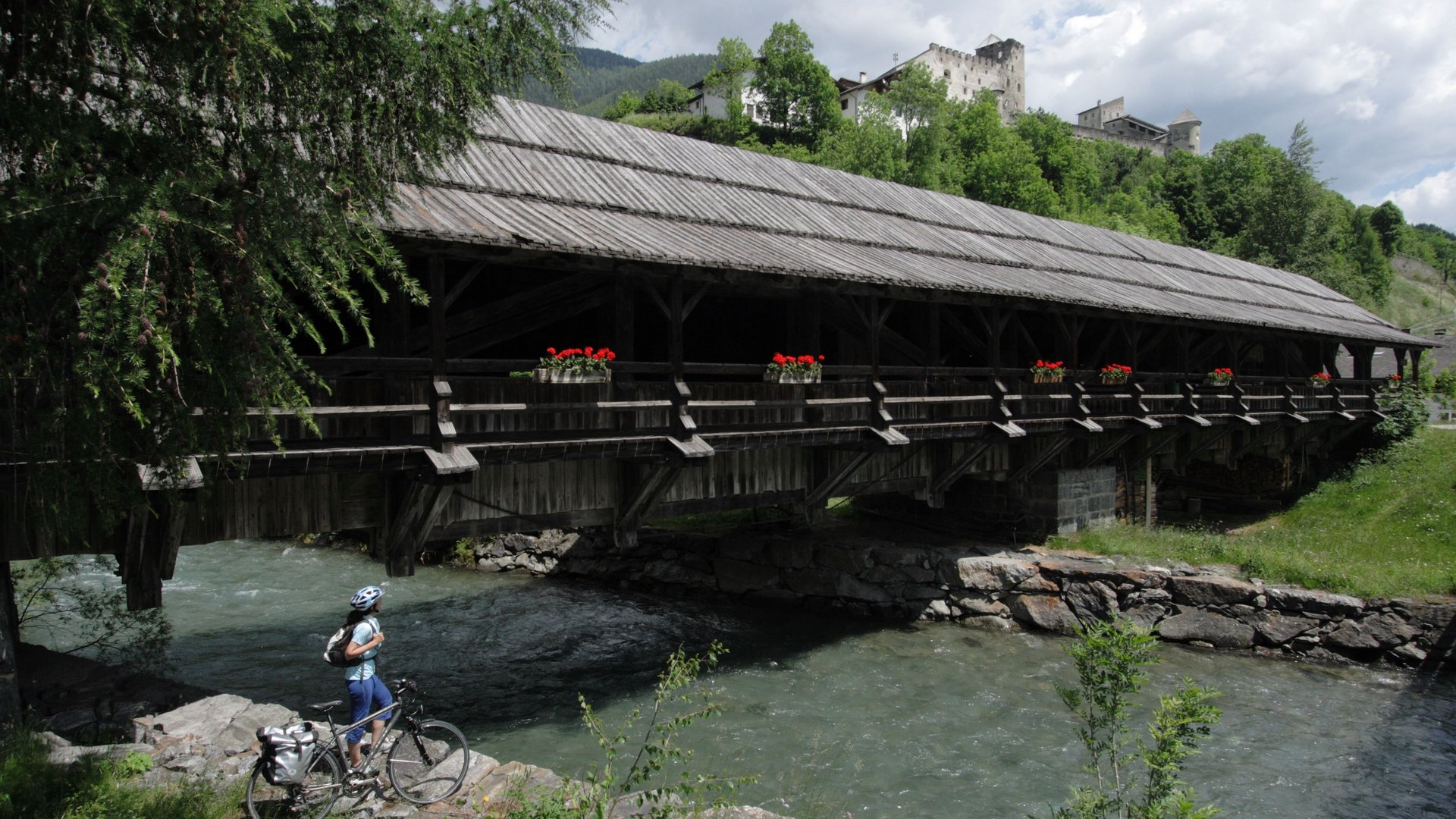 Historische Holzbrücke Überdachte Holzbrücke mit Blumen, umgeben von grüner Landschaft und fließendem Fluss.
