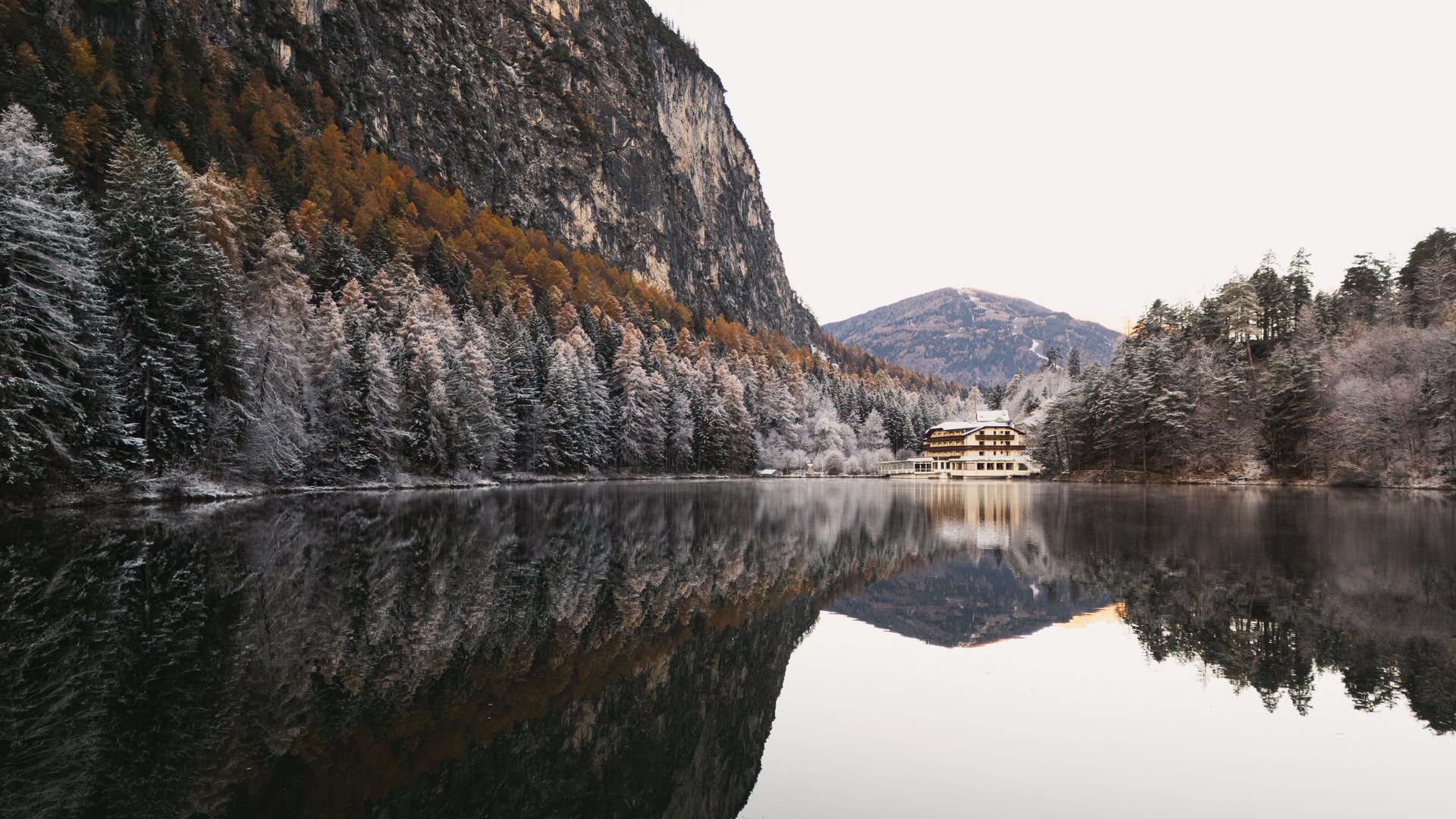 Winterzauber am See Hotel am verschneiten See, umgeben von schneebedeckten Bäumen und Bergen.