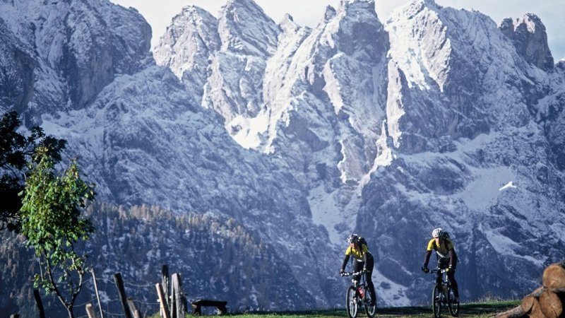 Zwei Radfahrer auf Bergpfad, umgeben von schneebedeckten Gipfeln und blauem Himmel.