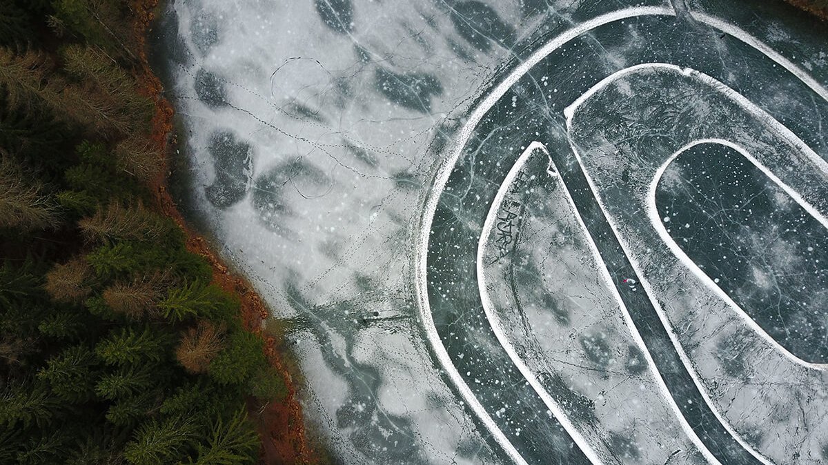 Luftaufnahme eines verschneiten Eislaufplatzes am Waldrand mit klaren Linien und Mustern.