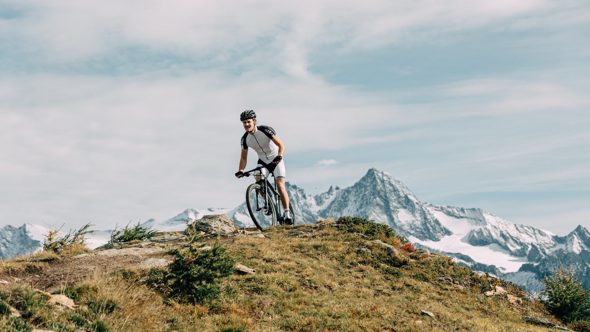 Radfahrer auf einem Bergpfad mit schneebedeckten Alpengipfeln im Hintergrund.