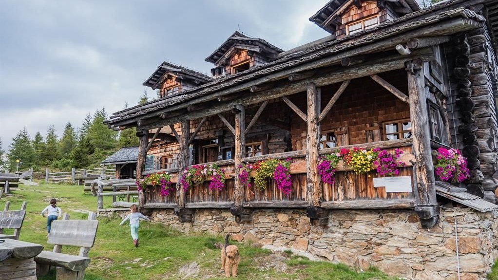 Traditionelle Berghütte Rustikale Berghütte mit Blumenschmuck, umgeben von grüner Natur und spielenden Kindern.